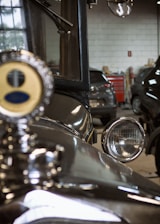 An old vintage car is parked in a dimly lit garage with various car workshop tools visible in the background. The car's headlight and radiator cap are highlighted, reflecting its shiny, polished surface.