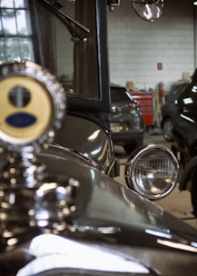 An old vintage car is parked in a dimly lit garage with various car workshop tools visible in the background. The car's headlight and radiator cap are highlighted, reflecting its shiny, polished surface.