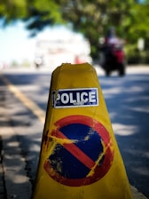 A yellow traffic cone with a 'Police' label and a faded red and blue no-parking symbol is positioned on the edge of a road. In the background, the blurred image of a motorcyclist and greenery is visible, suggesting a street setting.