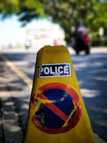 A yellow traffic cone with a 'Police' label and a faded red and blue no-parking symbol is positioned on the edge of a road. In the background, the blurred image of a motorcyclist and greenery is visible, suggesting a street setting.