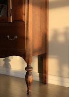 A wooden piece of furniture with intricate carvings and a rich brown finish is bathed in warm sunlight. The shadow of the furniture leg is cast against the cream-colored wall, highlighting the elegant, turned design of the leg.