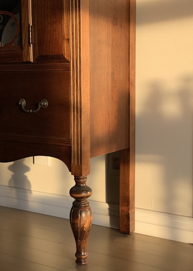 A wooden piece of furniture with intricate carvings and a rich brown finish is bathed in warm sunlight. The shadow of the furniture leg is cast against the cream-colored wall, highlighting the elegant, turned design of the leg.
