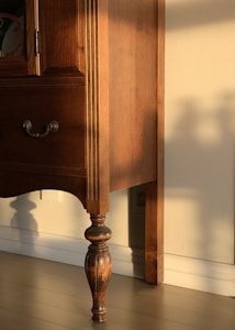 A wooden piece of furniture with intricate carvings and a rich brown finish is bathed in warm sunlight. The shadow of the furniture leg is cast against the cream-colored wall, highlighting the elegant, turned design of the leg.