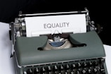 A vintage typewriter holds a sheet of white paper with the word 'EQUALITY' typed on it. The typewriter has a dark green body and black keys. The scene is set on a white and black background, giving it a minimalist yet impactful appearance.