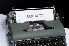 A vintage typewriter holds a sheet of white paper with the word 'EQUALITY' typed on it. The typewriter has a dark green body and black keys. The scene is set on a white and black background, giving it a minimalist yet impactful appearance.