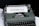 An old-fashioned typewriter with a piece of white paper inserted, displaying the word 'MOTHERHOOD' typed in bold letters. The typewriter has a vintage design with dark keys and a greenish-gray body.