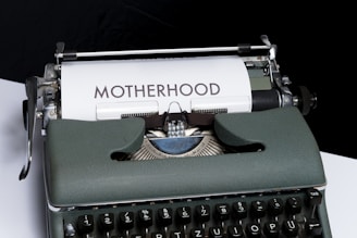 An old-fashioned typewriter with a piece of white paper inserted, displaying the word 'MOTHERHOOD' typed in bold letters. The typewriter has a vintage design with dark keys and a greenish-gray body.