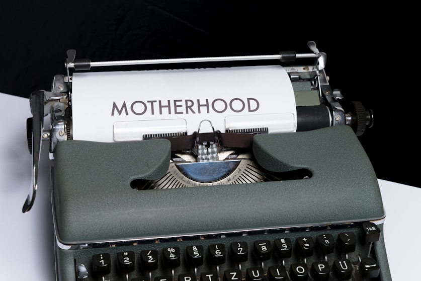 An old-fashioned typewriter with a piece of white paper inserted, displaying the word 'MOTHERHOOD' typed in bold letters. The typewriter has a vintage design with dark keys and a greenish-gray body.