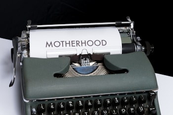 An old-fashioned typewriter with a piece of white paper inserted, displaying the word 'MOTHERHOOD' typed in bold letters. The typewriter has a vintage design with dark keys and a greenish-gray body.