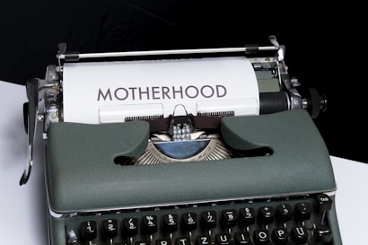 An old-fashioned typewriter with a piece of white paper inserted, displaying the word 'MOTHERHOOD' typed in bold letters. The typewriter has a vintage design with dark keys and a greenish-gray body.