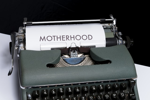 An old-fashioned typewriter with a piece of white paper inserted, displaying the word 'MOTHERHOOD' typed in bold letters. The typewriter has a vintage design with dark keys and a greenish-gray body.