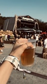 A hand is holding a plastic cup filled with beer at an outdoor festival. In the background, a stage with a sign and a gathering of people enjoying the event can be seen. Some are seated or standing near hay bales, and a sunny sky enhances the vibrant atmosphere.
