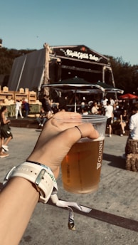A hand is holding a plastic cup filled with beer at an outdoor festival. In the background, a stage with a sign and a gathering of people enjoying the event can be seen. Some are seated or standing near hay bales, and a sunny sky enhances the vibrant atmosphere.