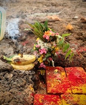 A colorful ritual setup is arranged on the ground. Fresh flowers, including pink and yellow blossoms, are placed next to green leaves. Nearby, several bananas rest on the soil with incense sticks burning, emitting smoke. Red and yellow powders are sprinkled across bricks laid in an orderly fashion.