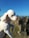 A small, white poodle is being held up with a clear blue sky and rocky landscape in the background. The dog's fur is neatly groomed, and its eyes are closed as it faces the sunlight.