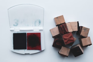 A set of wooden rubber stamps arranged in a circular pattern next to an open ink pad containing two colors, black and red. The stamps seem to be used for printing letters or symbols.
