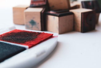 A set of wooden blocks, some with visible ink stamps, are arranged on a white surface. The foreground features open red and black ink pads, suggesting a crafting or stamping activity.