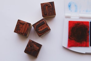 Four wooden stamps with letters in reverse are arranged on a white surface next to an open ink pad. The stamps appear worn with a rustic texture, and the ink pad has red and blue sections with visible marks of use.