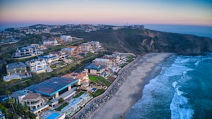 A coastal scene featuring luxury homes along a hillside, overlooking a sandy beach with calm waves. The sky is a gradient of pastel colors indicating dawn or dusk.