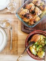A dining table setting with a glass baking dish containing seasoned and cooked chicken pieces, likely with herbs, placed on a wooden board. Beside it, a decorative bowl holds a serving of potatoes and broccoli. The table is set with woven placemats, plates, cutlery, and glasses of water.