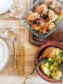 A dining table setting with a glass baking dish containing seasoned and cooked chicken pieces, likely with herbs, placed on a wooden board. Beside it, a decorative bowl holds a serving of potatoes and broccoli. The table is set with woven placemats, plates, cutlery, and glasses of water.