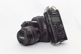 A digital SLR camera and a digital audio recorder are placed together on a light surface. The camera is equipped with a lens and is branded Canon, while the audio recorder is a Tascam DR-40 model. The camera has a sleek black body with several buttons and controls visible. The audio recorder features various control buttons and a small screen.