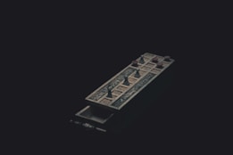 A dark image featuring an ornate, ancient-looking game board with small figurines resembling pawns placed on it. The board has intricate designs and is positioned diagonally across the frame. The overall lighting is dim, casting shadows that create a mysterious atmosphere.