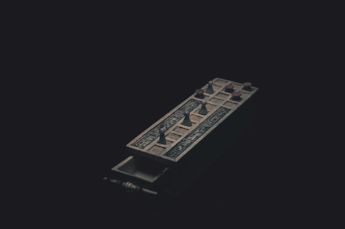 A dark image featuring an ornate, ancient-looking game board with small figurines resembling pawns placed on it. The board has intricate designs and is positioned diagonally across the frame. The overall lighting is dim, casting shadows that create a mysterious atmosphere.