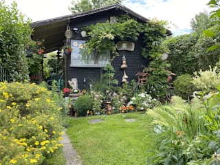 A charming wooden garden shed is surrounded by lush greenery and a variety of colorful flowers. Vines are growing around the structure, and there are decorative elements such as hanging gourds and garden ornaments. The lawn is neatly maintained, and the atmosphere is serene and inviting.