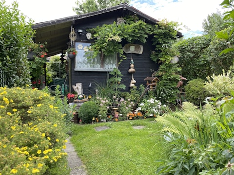 A charming wooden garden shed is surrounded by lush greenery and a variety of colorful flowers. Vines are growing around the structure, and there are decorative elements such as hanging gourds and garden ornaments. The lawn is neatly maintained, and the atmosphere is serene and inviting.