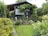 A charming wooden garden shed is surrounded by lush greenery and a variety of colorful flowers. Vines are growing around the structure, and there are decorative elements such as hanging gourds and garden ornaments. The lawn is neatly maintained, and the atmosphere is serene and inviting.