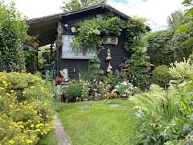 A charming wooden garden shed is surrounded by lush greenery and a variety of colorful flowers. Vines are growing around the structure, and there are decorative elements such as hanging gourds and garden ornaments. The lawn is neatly maintained, and the atmosphere is serene and inviting.