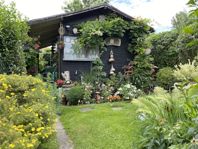 A charming wooden garden shed is surrounded by lush greenery and a variety of colorful flowers. Vines are growing around the structure, and there are decorative elements such as hanging gourds and garden ornaments. The lawn is neatly maintained, and the atmosphere is serene and inviting.