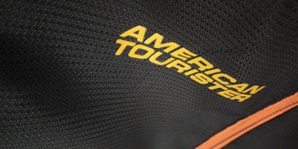 The close-up view captures a textured black fabric with the words 'American Tourister' printed in bold yellow letters. A contrasting orange line runs diagonally along the fabric, potentially indicating a zipper or trim on a bag or suitcase.