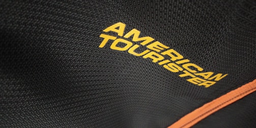 The close-up view captures a textured black fabric with the words 'American Tourister' printed in bold yellow letters. A contrasting orange line runs diagonally along the fabric, potentially indicating a zipper or trim on a bag or suitcase.