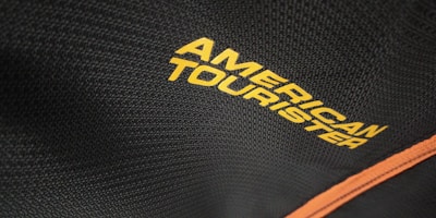 The close-up view captures a textured black fabric with the words 'American Tourister' printed in bold yellow letters. A contrasting orange line runs diagonally along the fabric, potentially indicating a zipper or trim on a bag or suitcase.