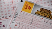 A close-up image of lottery tickets featuring crossed-out numbers, brand logos, and various numbers in a grid format. The tickets have sections for different games, and numbers are marked with black crosses.