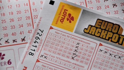 A close-up image of lottery tickets featuring crossed-out numbers, brand logos, and various numbers in a grid format. The tickets have sections for different games, and numbers are marked with black crosses.
