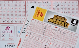 Three lottery tickets are displayed, including Eurojackpot and Super 6. Various numbers are marked on the tickets, with some crossed out and others checked. The Eurojackpot ticket is partially visible with the logo prominently displayed, and a ticket number is visible on one of the tickets.