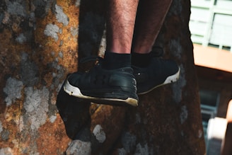 A person wearing black sneakers is climbing a rock surface, with their legs visible. The rock has a textured appearance with patches of brown and grey. Sunlight casts shadows, creating a contrast between light and dark areas. Part of a building with green and orange colors is visible in the background.