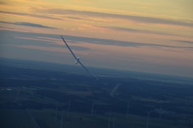 A glider gracefully soars through the sky at sunset, with its wings perfectly aligned to catch the light. Below, a landscape dotted with wind turbines stretches into the distance, creating a sense of tranquility and harmony with nature.
