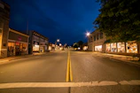 A quiet small town street at dusk, warm lights glowing from storefronts, inviting connection.