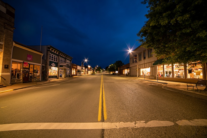 A small town street at night, lined with quaint shops that are illuminated by warm lighting. The sky is a deep blue, contrasting with the soft yellow glow from the streetlights. The street is empty, creating a calm and peaceful atmosphere.