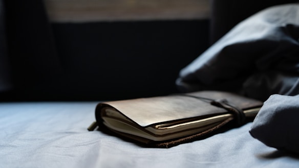 A leather-bound journal rests on a soft, crumpled bedspread in a dimly lit room. The journal is slightly open, and the ambient lighting casts soft shadows around it, creating a calm and intimate atmosphere.