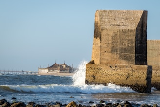 A large concrete structure juts into the sea with waves crashing against it. In the background, there is a pier with a building that has pointed rooftops. The scene conveys a sense of coastal architecture and natural interaction with the ocean.