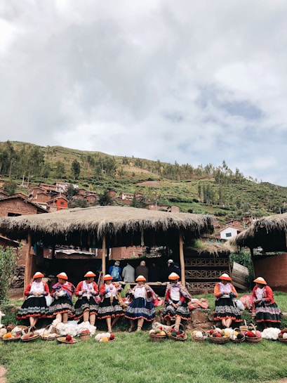 A group of women wearing traditional clothing with bright colors and intricate patterns are seated outside, possibly in a rural setting. They appear to be engaging in activities such as knitting or weaving. In front of them are baskets filled with yarn or textiles. The background features grassy hills and a cloudy sky, along with a few thatched-roof structures.