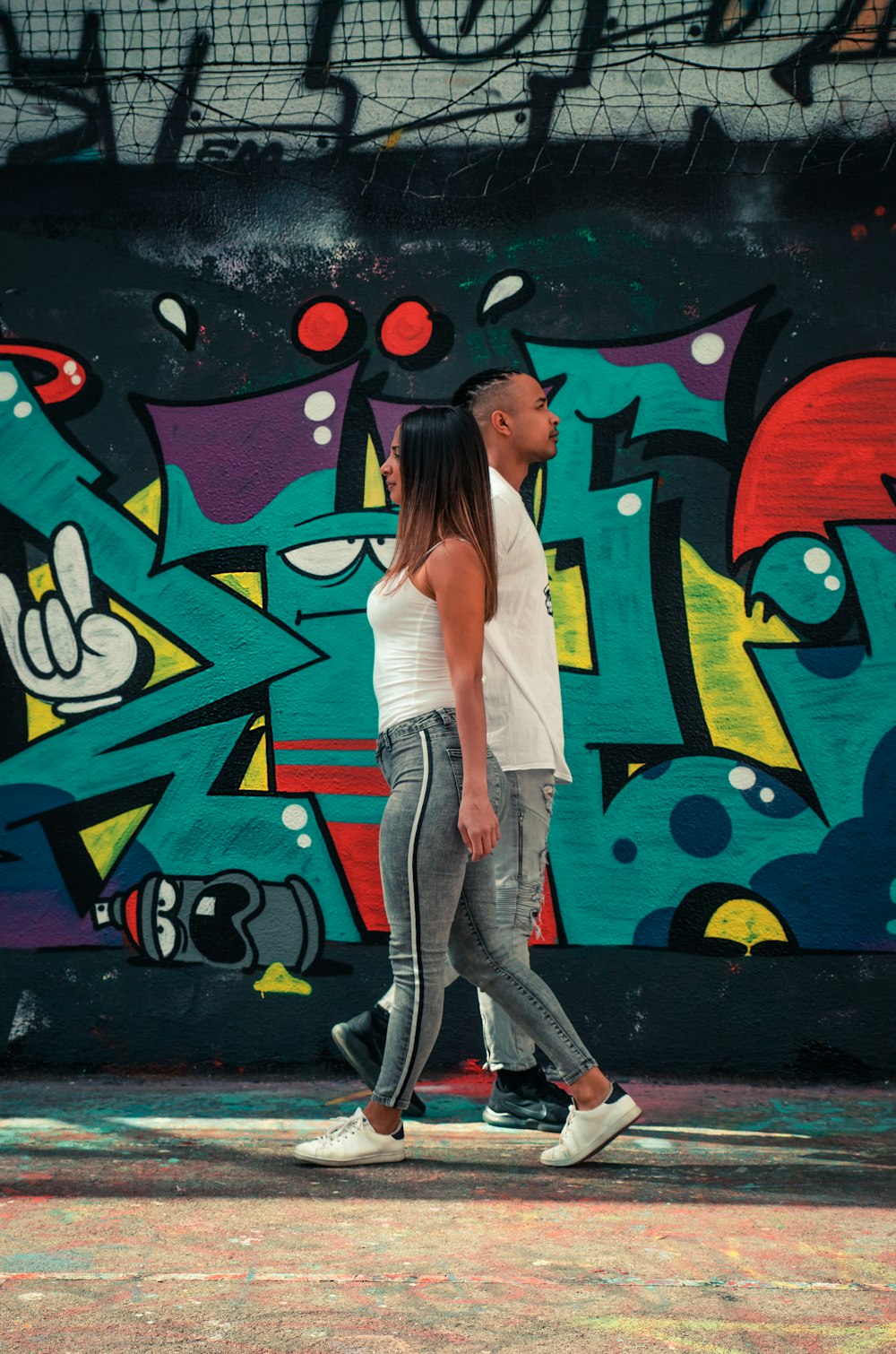 Mujer con camiseta blanca sin mangas y jeans de mezclilla grises de pie junto a la pared de graffiti