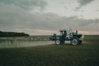 A large agricultural machine is spraying crops in a vast, open field. It is overcast with a cloudy sky, and the landscape is expansive with distant tree lines.