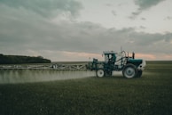 A large agricultural machine is spraying crops in a vast, open field. It is overcast with a cloudy sky, and the landscape is expansive with distant tree lines.