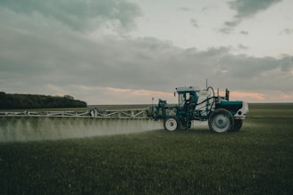 A large agricultural machine is spraying crops in a vast, open field. It is overcast with a cloudy sky, and the landscape is expansive with distant tree lines.